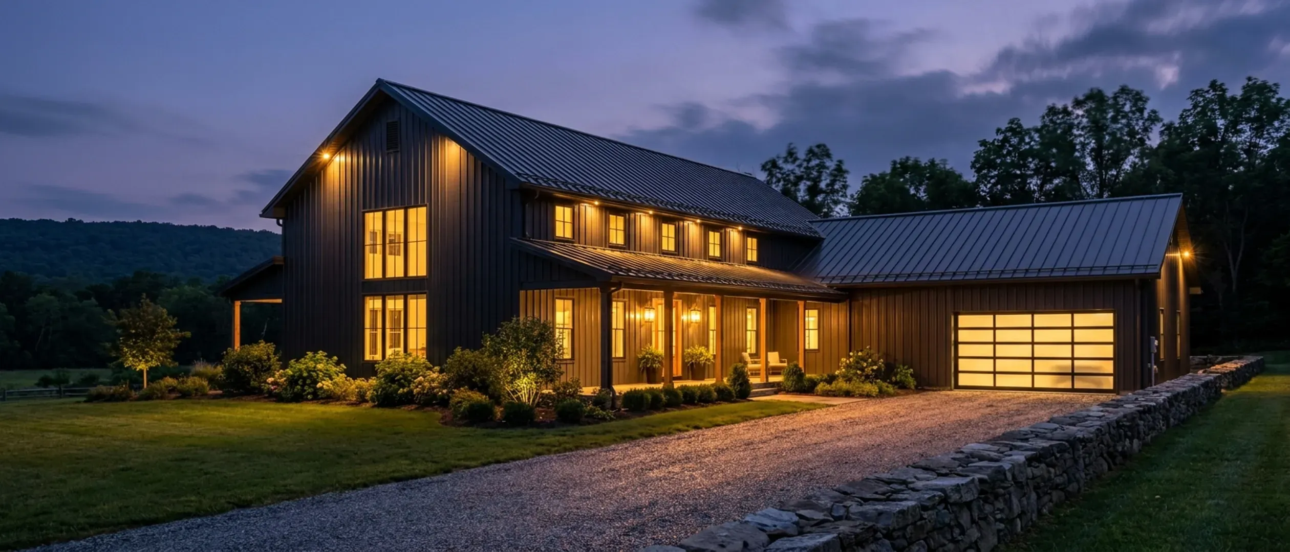 A barndominium at dusk with warm light glowing from every window and exterior lighting along the porch and roofline against a deep blue twilight sky.